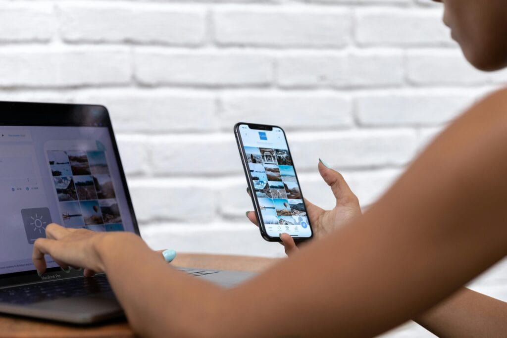 Person holding a smartphone and selecting images on a laptop, with a white brick wall in the background. | Tsunami Reach