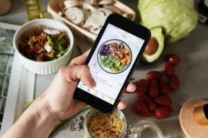 Person holding a phone showing a food photo; fresh vegetables and salad ingredients are on the table. | Tsunami Reach