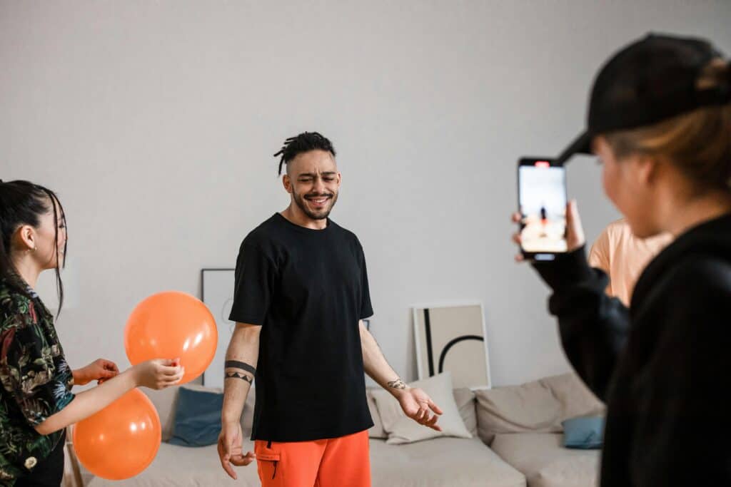 Man smiling and posing while a person takes his photo and another holds orange balloons in a living room. | Tsunami Reach