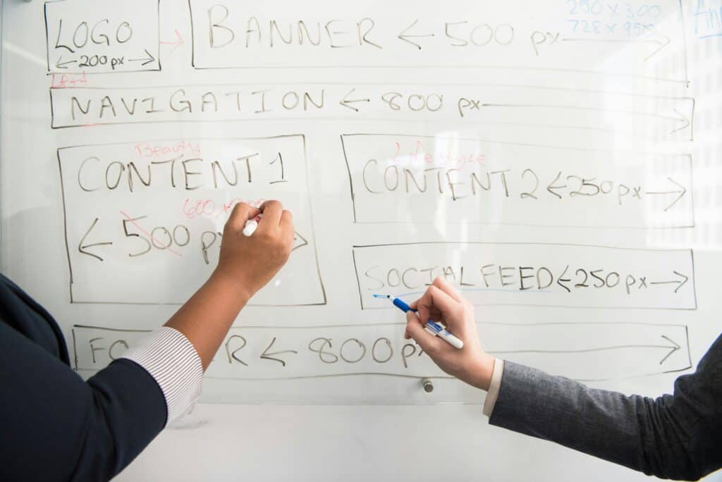 Two people write a website wireframe with sections and pixel sizes on a whiteboard using markers. | Tsunami Reach