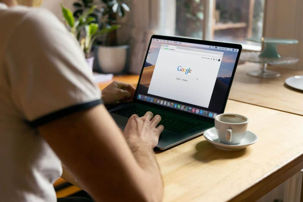 Person using a laptop with Google open on screen, seated at a wooden table with a cup of coffee and plants in the background. | Tsunami Reach