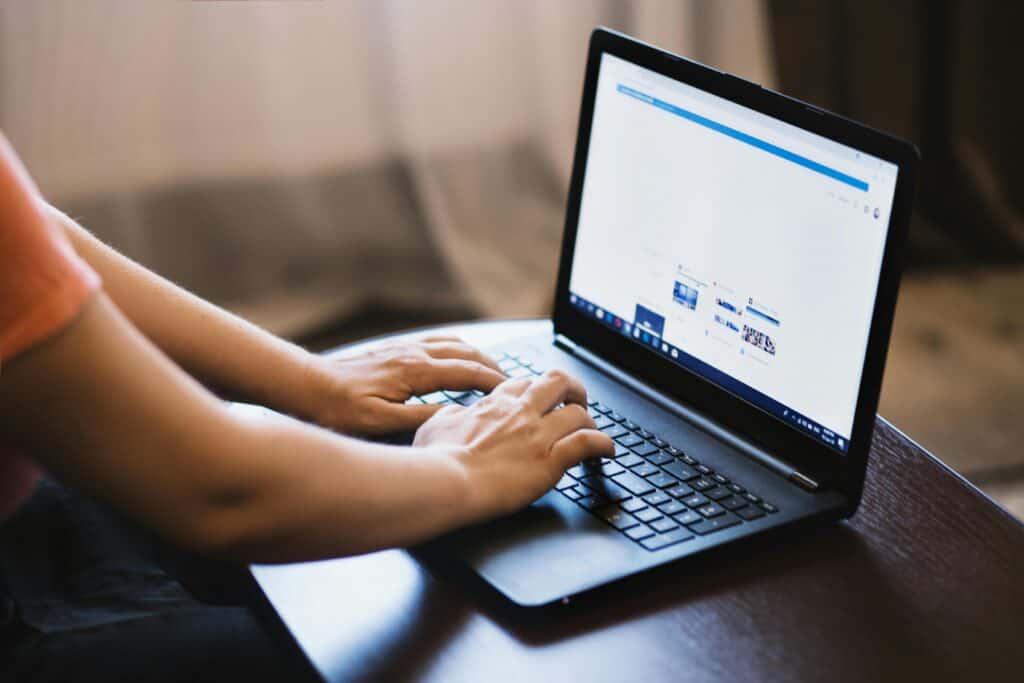 Person typing on a black laptop at a table, browsing the internet with a bright window in the background. | Tsunami Reach