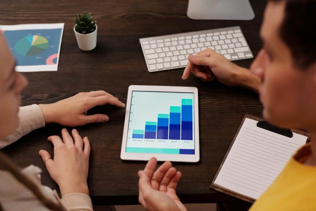 Two people discuss a bar graph on a tablet at a desk with a keyboard, clipboard, and a small plant. | Tsunami Reach