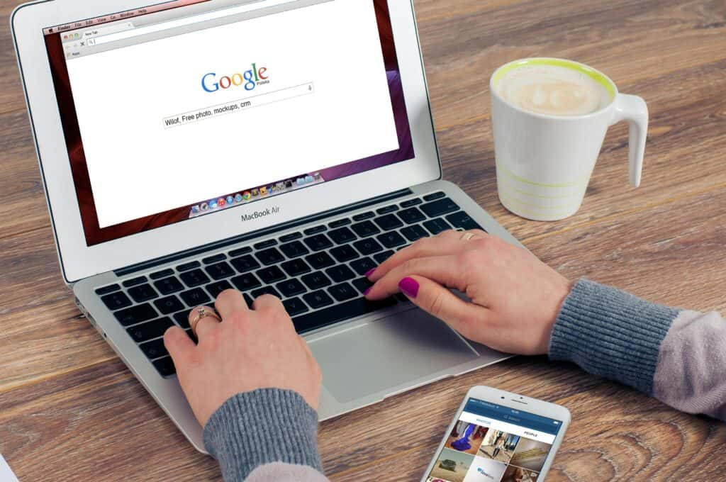 Person typing on a MacBook Air with Google open, next to a coffee and a smartphone on a wooden desk. | Tsunami Reach