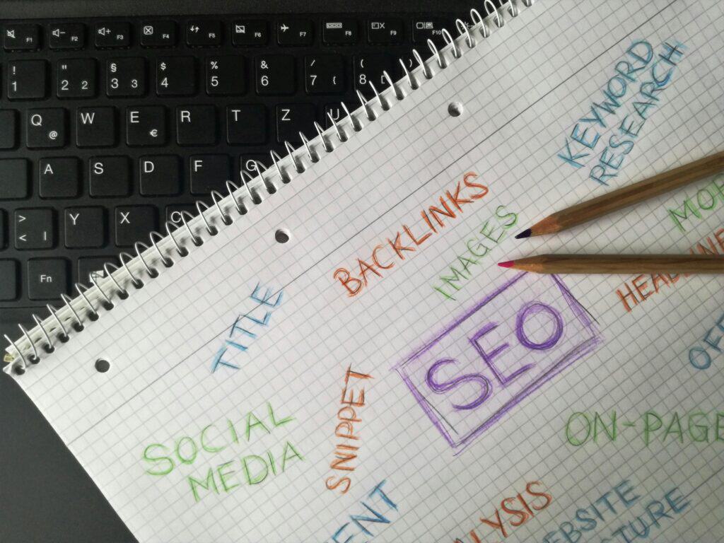 Notebook with SEO-related terms written in colorful text, placed on a laptop keyboard with two pencils on top. | Tsunami Reach