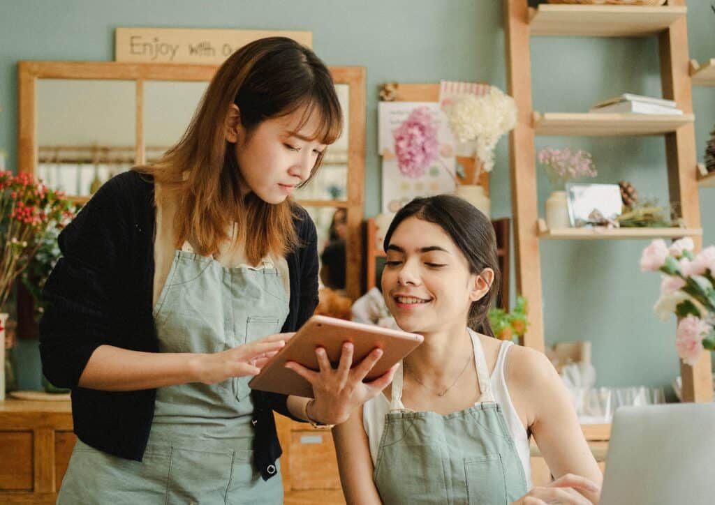Two women wearing aprons look at a tablet together and smile in a cozy, flower-filled shop. | Tsunami Reach