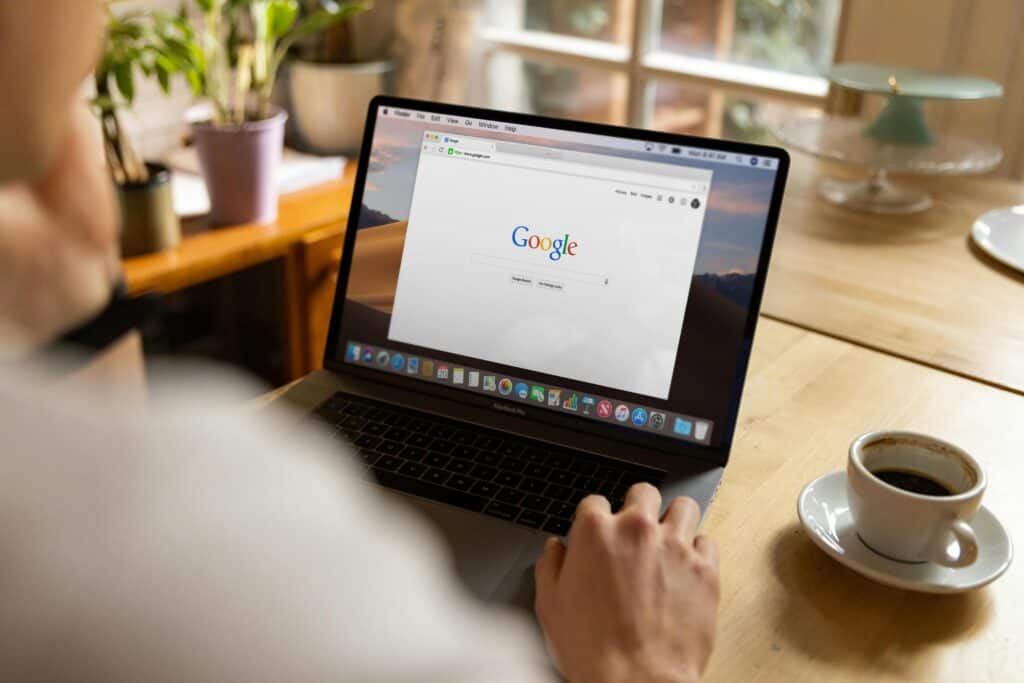 Person using a laptop with the Google homepage open, sitting at a wooden table with coffee and plants nearby. | Tsunami Reach
