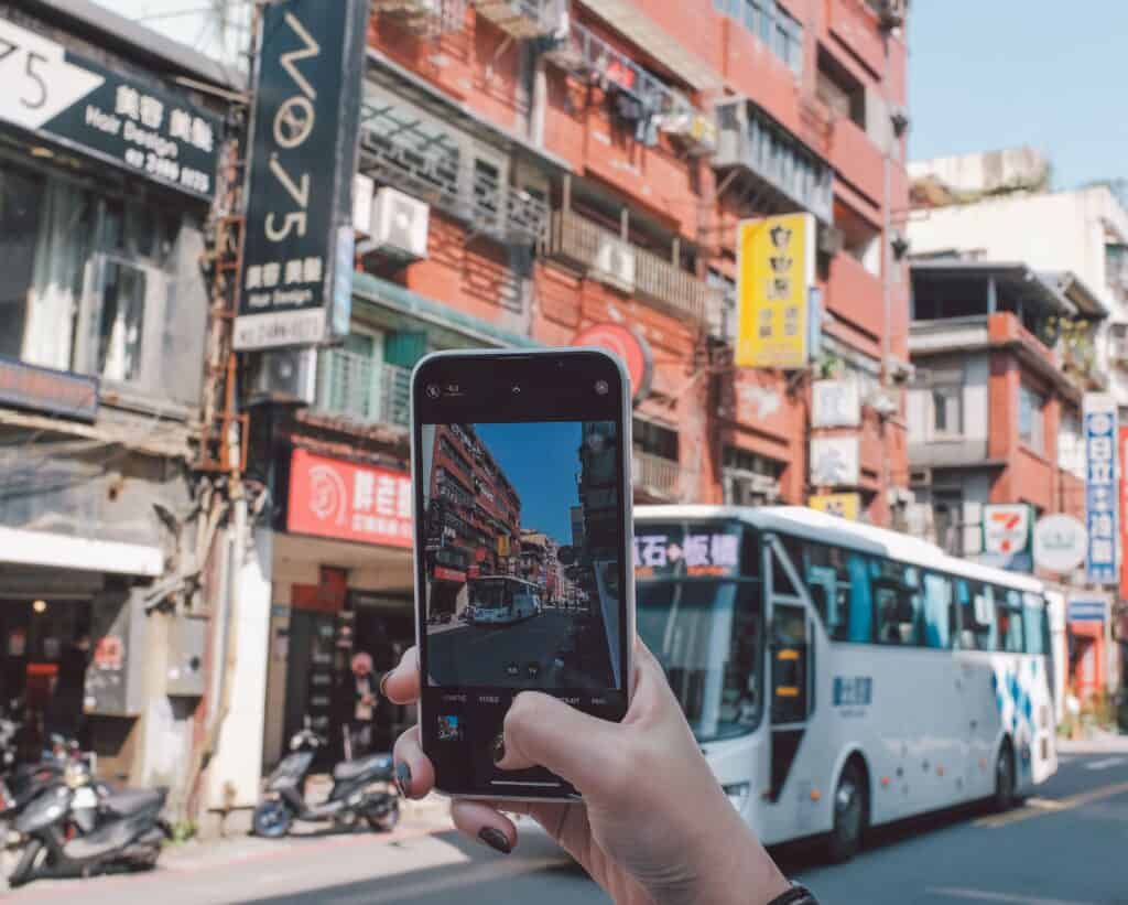 A person takes a photo of a busy city street with shops, signs, and a white bus passing by. | Tsunami Reach