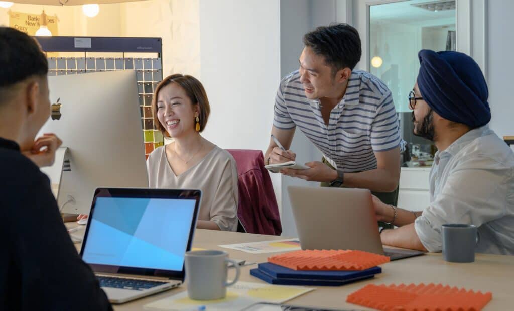 Four people in an office collaborate on digital marketing around a desk with laptops, smiling and discussing ideas. | Tsunami Reach
