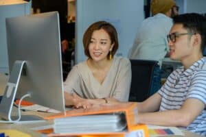 Two people sit at a desk, looking at a computer monitor and discussing work in an office setting. | Tsunami Reach