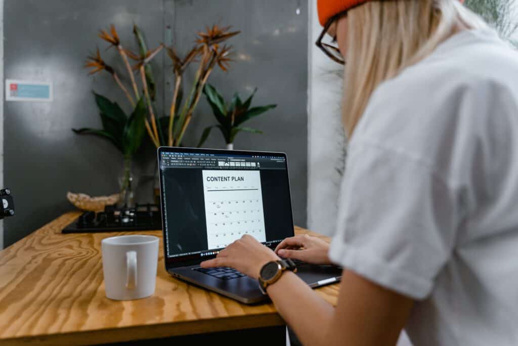 Person wearing glasses and an orange beanie develops a content marketing plan on a laptop at a wooden table with a mug. | Tsunami Reach