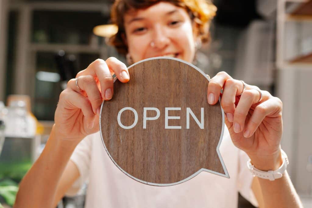 Smiling person holding a round wooden sign that says "OPEN" in white letters. | Tsunami Reach