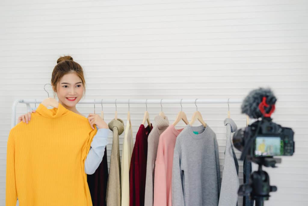 Woman holding a yellow sweater by a rack, filming content on digital marketing for online stores with a tripod camera. | Tsunami Reach