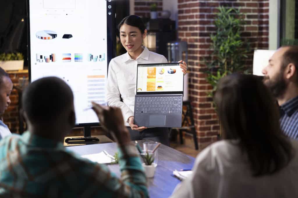 Woman presenting data on a laptop to colleagues in a meeting room with a digital chart displayed on a screen. | Tsunami Reach