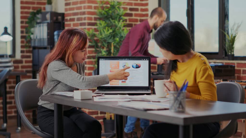 Two women discuss graphs on a laptop at a modern office, planning strategies for an SEO agency Singapore project. | Tsunami Reach