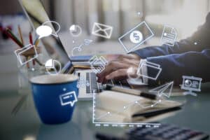 Person typing on laptop with digital business icons, coffee cup, notebook, and graph overlay on a desk. | Tsunami Reach