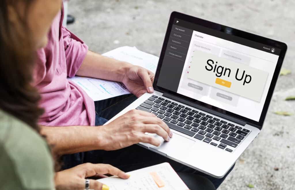 Two people sit outdoors. One uses a laptop showing a large “Sign Up” button on the screen. | Tsunami Reach