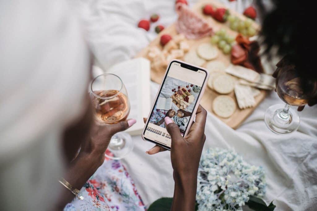 Two people at a picnic take a photo of a charcuterie board with a smartphone while holding glasses of rosé wine. | Tsunami Reach