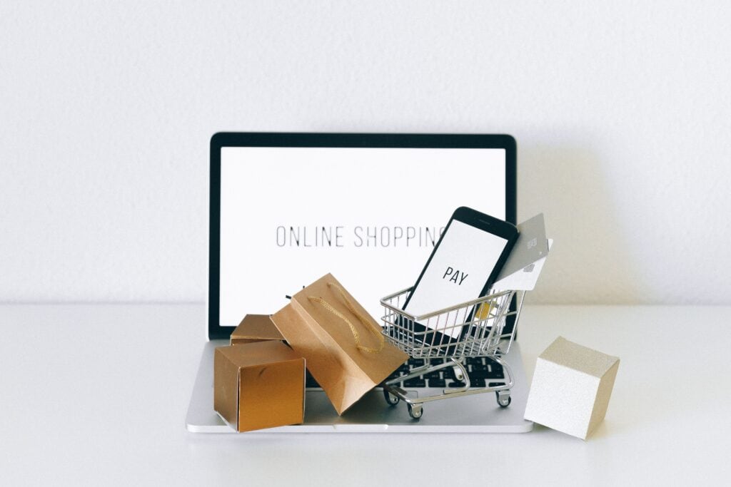 A laptop with "ONLINE SHOPPING" on screen, shopping bags, and a mini cart holding a phone and credit card in front. | Tsunami Reach