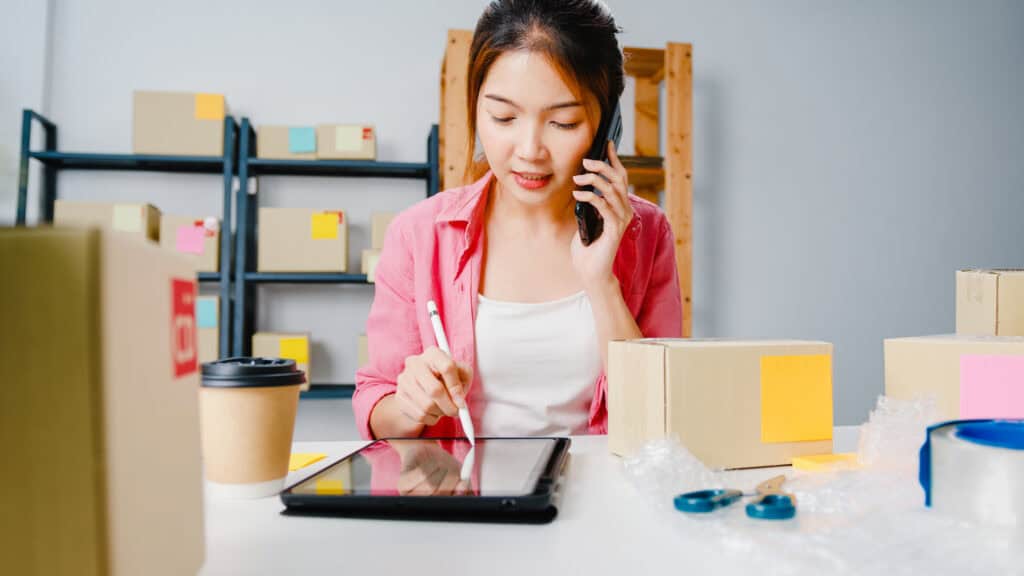 Woman talks on phone and writes on tablet at desk with boxes, planning digital marketing for online stores. | Tsunami Reach