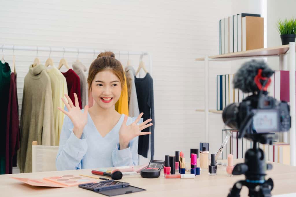 Woman filming makeup tutorial at table with cosmetics, smiling and gesturing, clothes hanging in background, camera visible. | Tsunami Reach