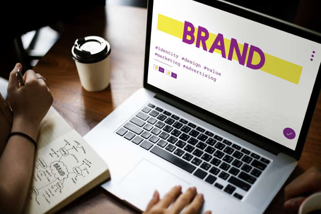 Two people work at a table with a laptop displaying “BRAND” and a notebook with handwritten notes. | Tsunami Reach