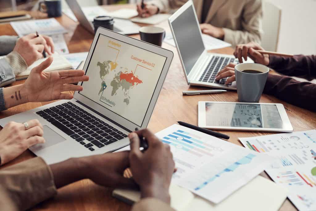 People working at a table with laptops, charts, and coffee, discussing a world map graph on a laptop screen. | Tsunami Reach