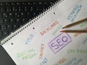 Spiral notebook with SEO-related words written in colorful pencils on a keyboard, two pencils cross on the page. | Tsunami Reach