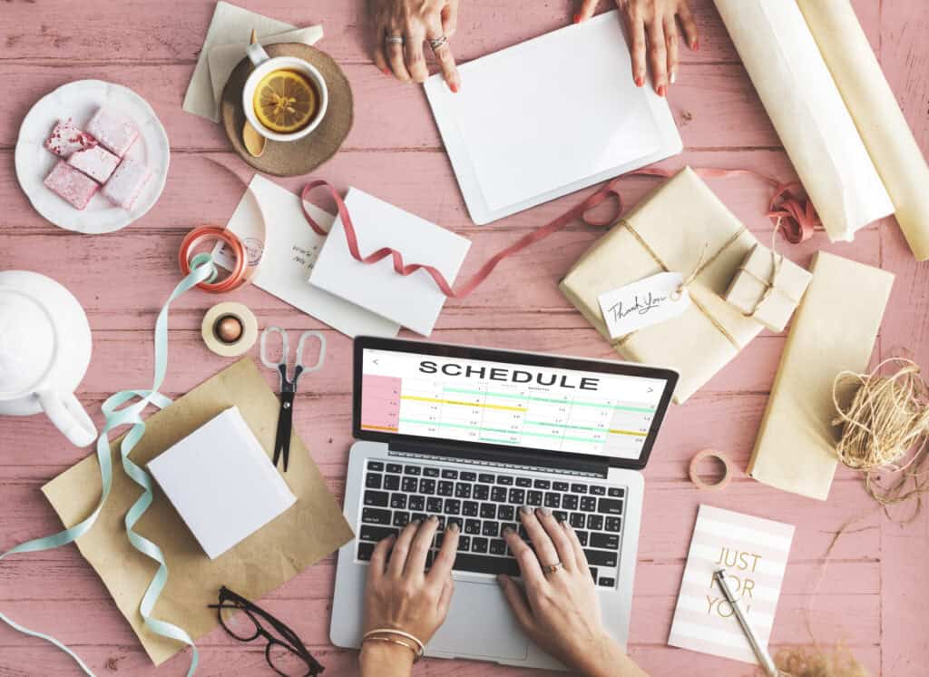 Hands using a laptop with a weekly content marketing schedule, gifts, ribbons, snacks, and a tablet on a pink table. | Tsunami Reach
