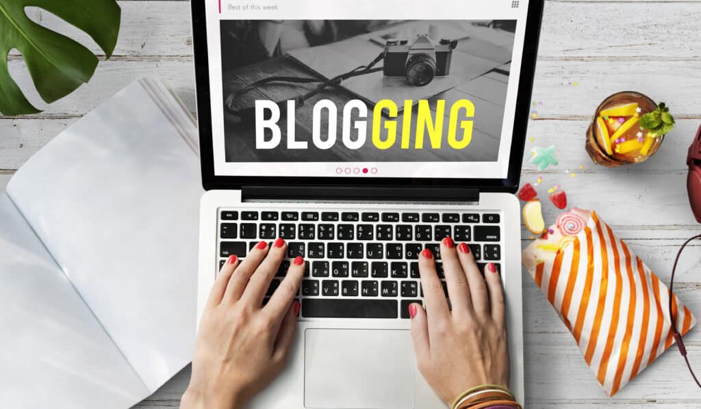 Hands typing on a laptop with “BLOGGING” on the screen, surrounded by notebook, camera, and snacks. | Tsunami Reach