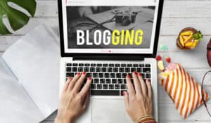 Hands typing on a laptop with “BLOGGING” on the screen, surrounded by notebook, camera, and snacks. | Tsunami Reach