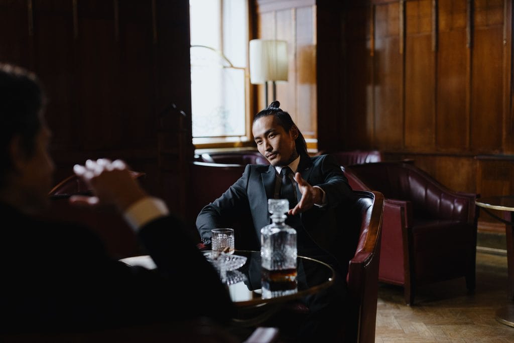 Two men in suits sit in leather chairs at a table with a glass decanter and a drink, talking in a wood-paneled room. | Tsunami Reach