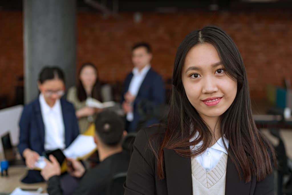 A young woman in professional attire smiles at the camera, with colleagues working in a blurred office background. | Tsunami Reach